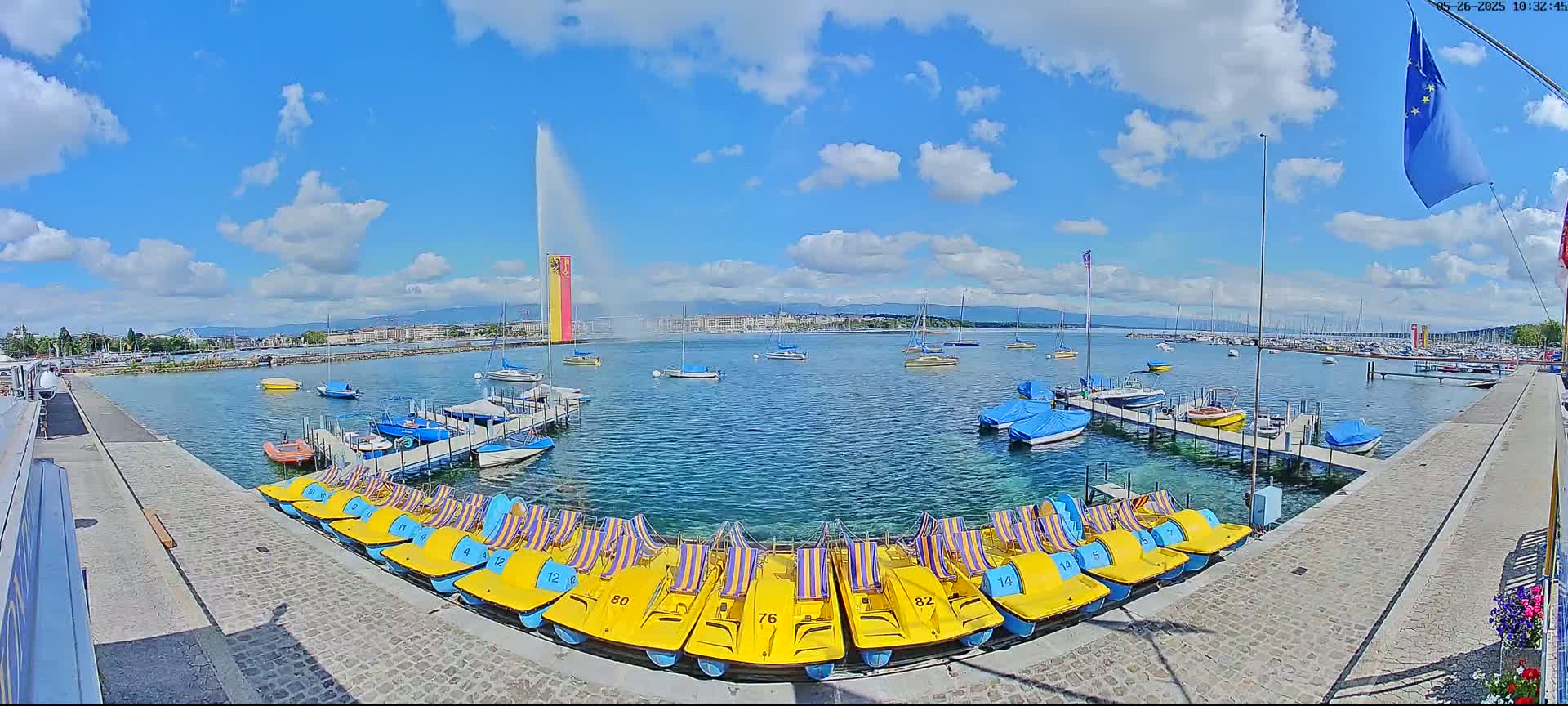 A large fountain sprays water into a sunny sky over a calm lake with numerous boats and pedal boats docked at a waterfront.