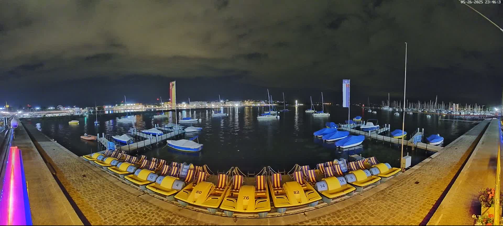 A nighttime view of a harbor filled with boats, under a cloudy sky, with numerous yellow pedal boats lined up on a dock.