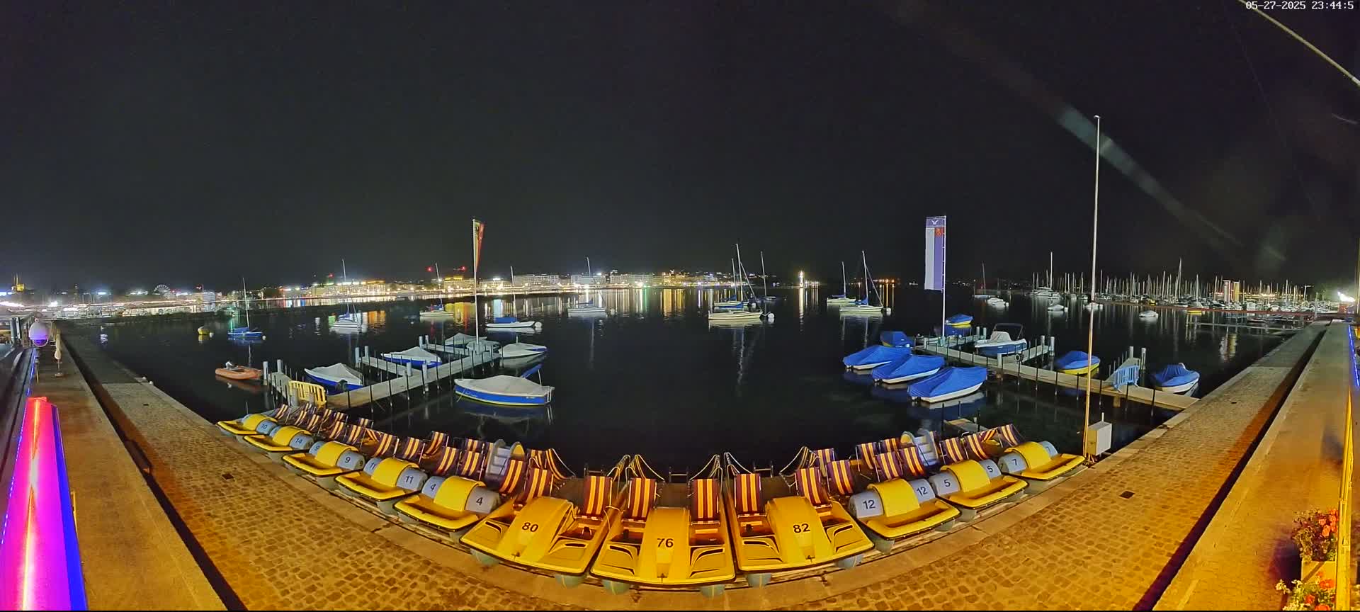 A nighttime view of a calm lake with docked boats and pedal boats lined up on a stone walkway, illuminated city lights in the background.