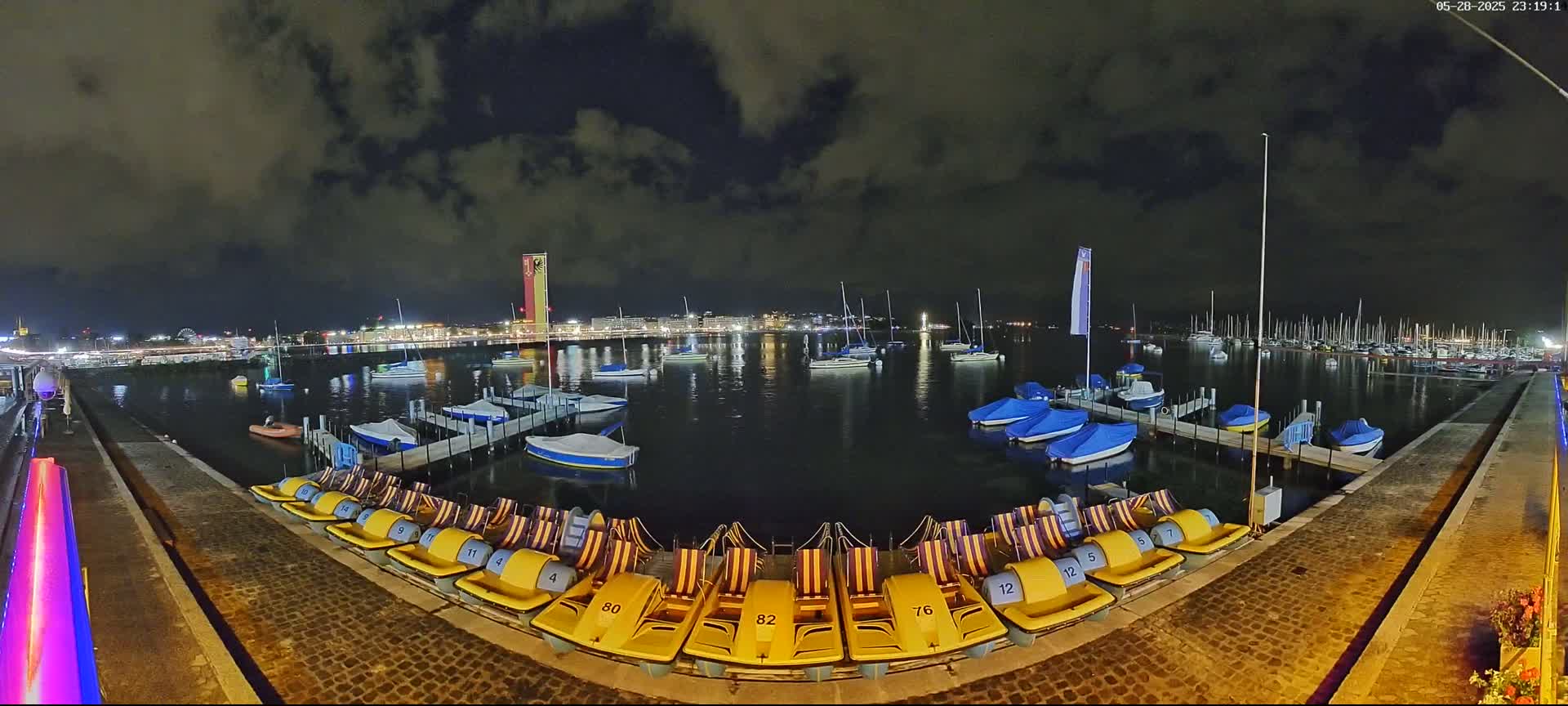 A nighttime view of a harbor filled with boats and docked pedal boats under a cloudy sky.