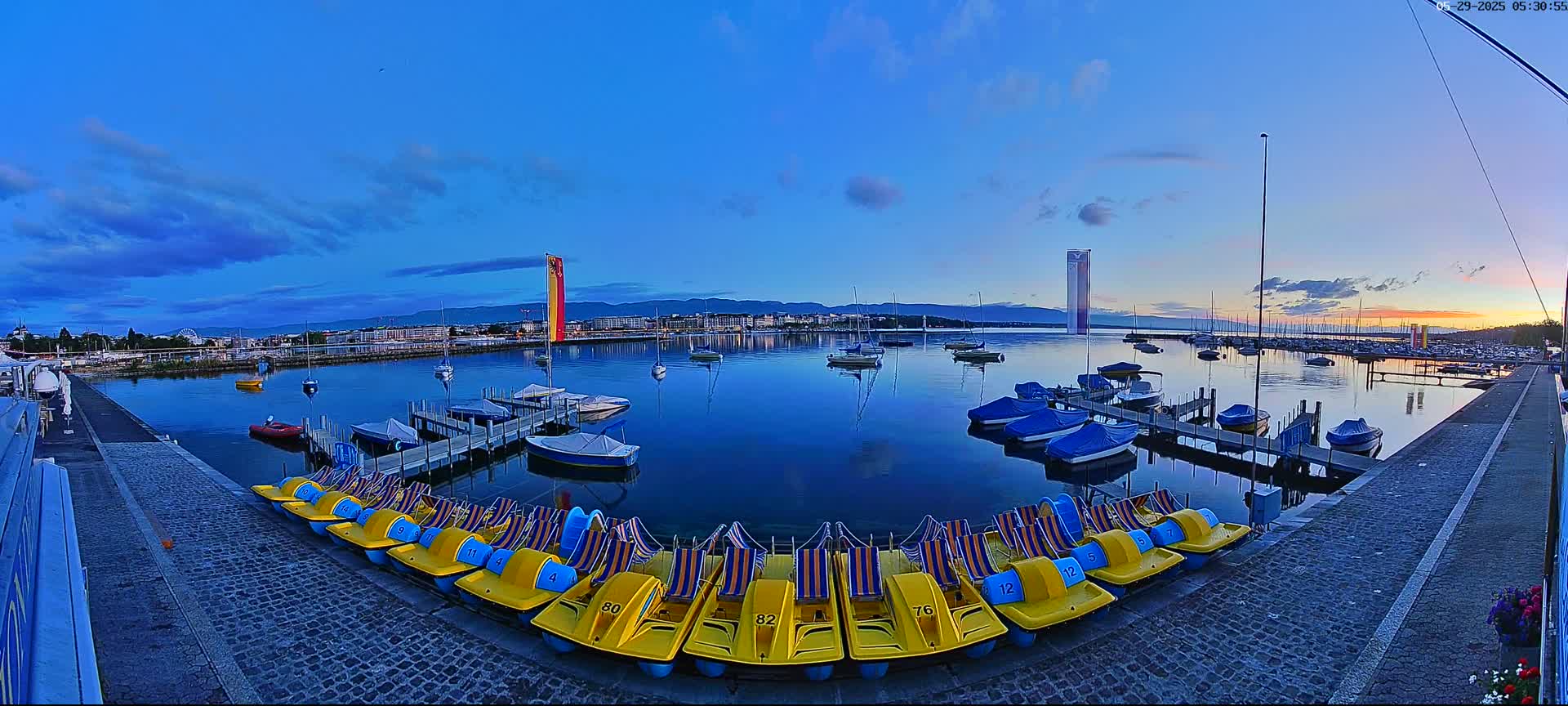 Many pedal boats are lined up on a stone dock next to a calm lake at sunset, with boats and a city skyline in the background.