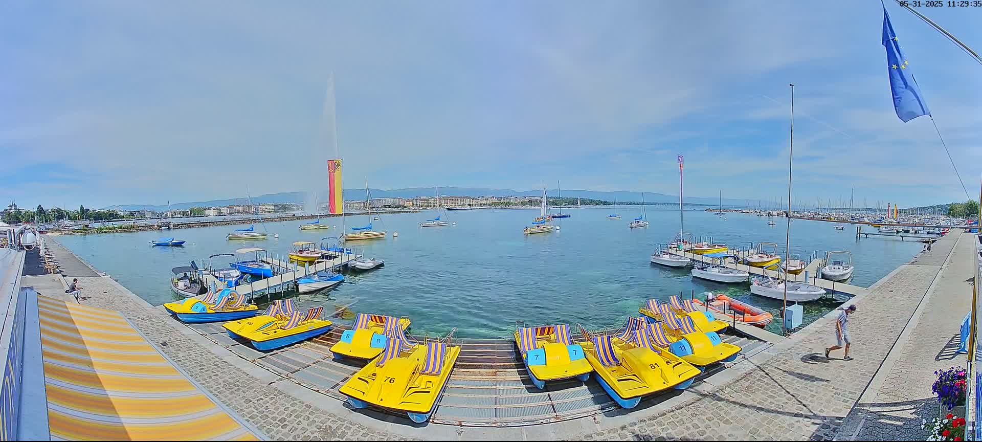 A harbor filled with boats and pedal boats on a sunny day, with a tall fountain in the background.