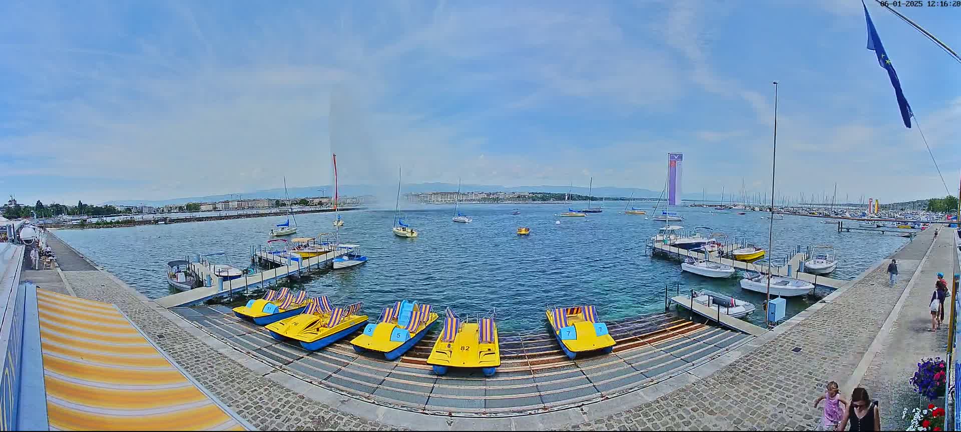 A harbor with numerous boats and pedal boats, a large water fountain in the distance, and a partly sunny sky.