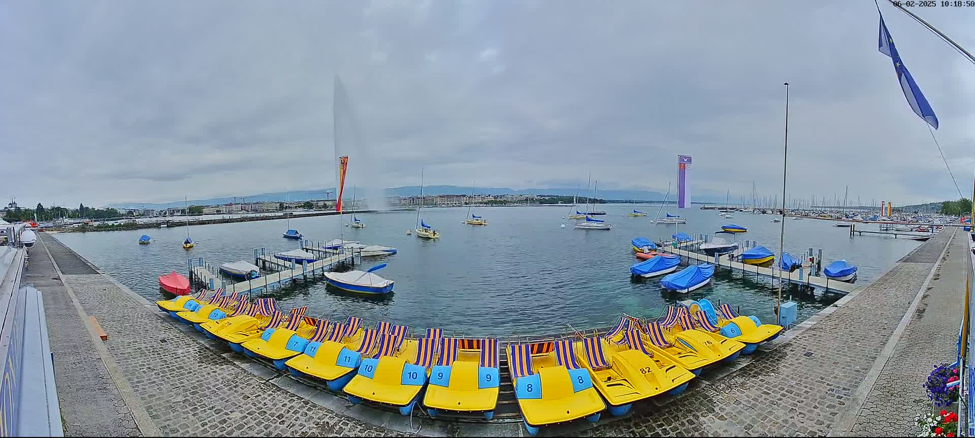 A large body of water with numerous boats and pedal boats docked, a tall fountain spraying water in the background, under an overcast sky.
