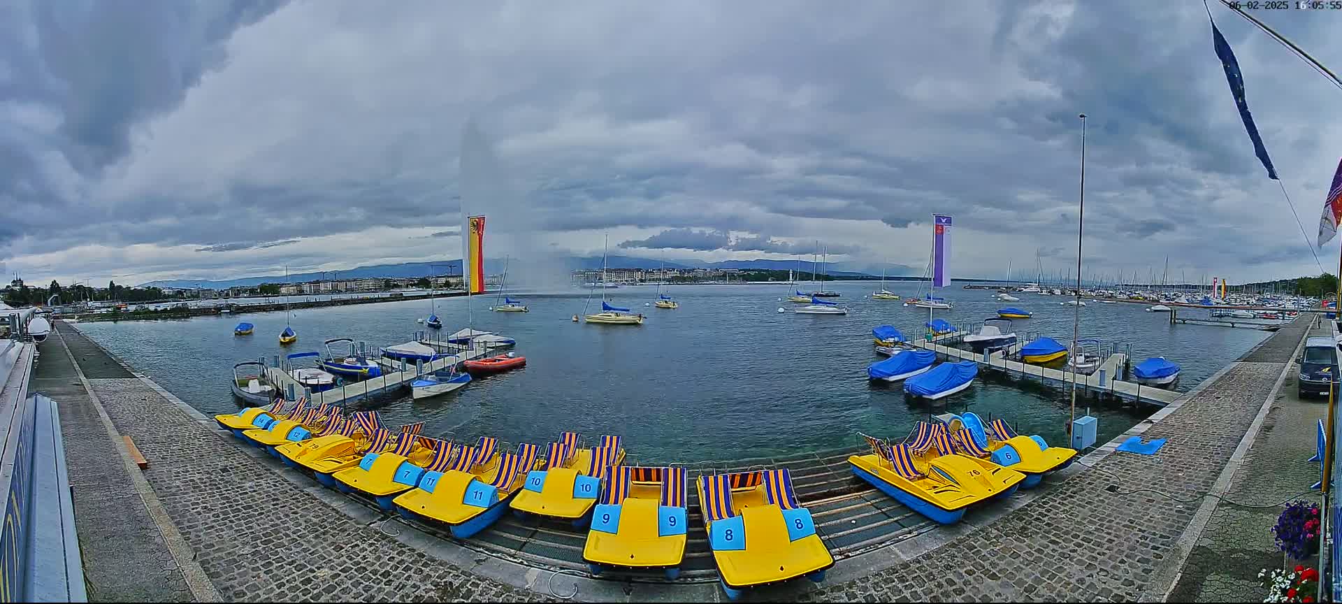 Under a cloudy sky, a harbor filled with docked boats and pedal boats is shown, with a large fountain and city skyline visible in the background.