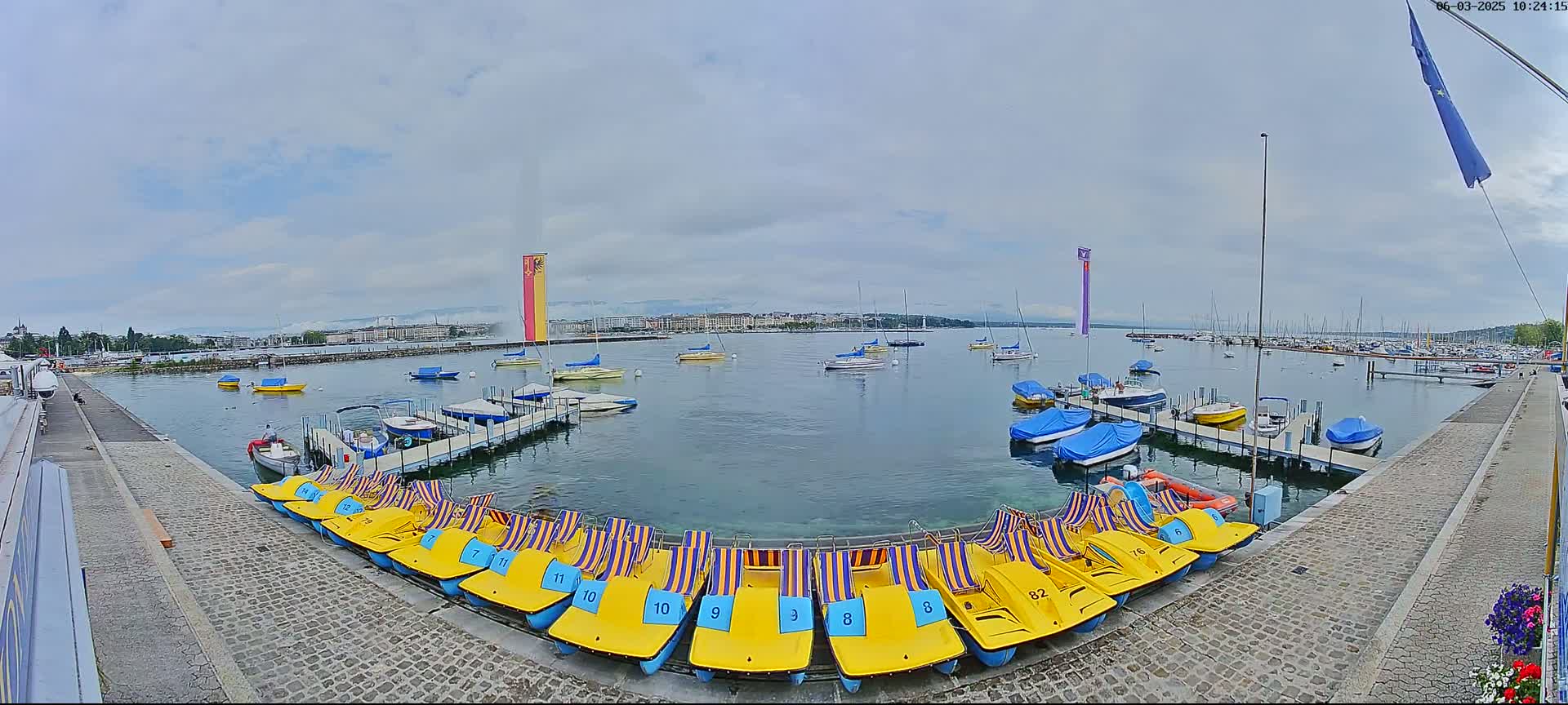 A calm lake with numerous boats and pedal boats docked at a stone pier under an overcast sky.