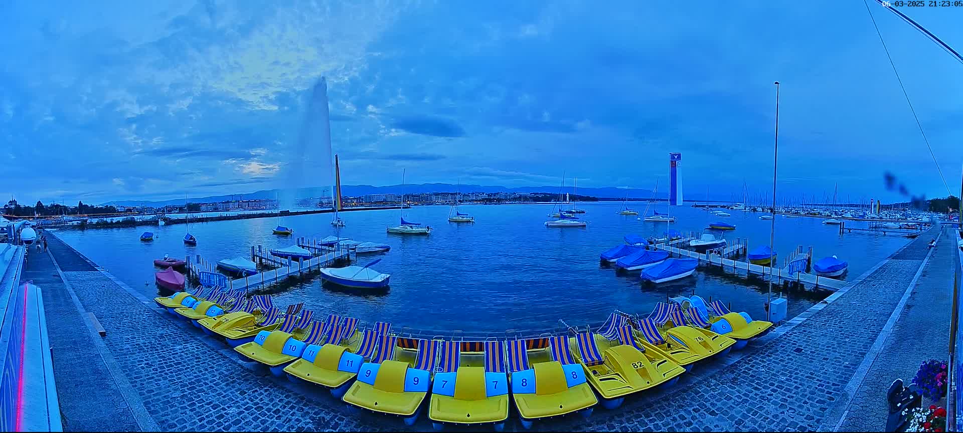 A body of water with numerous boats and pedal boats docked near a paved walkway, under a twilight sky with a large fountain in the background.