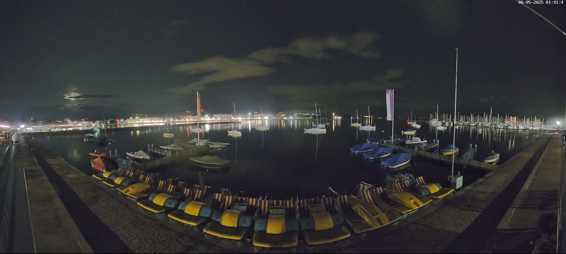 At night, under a partly cloudy sky, a calm body of water is dotted with sailboats and pedal boats moored at a dock, with a city skyline faintly visible in the distance.