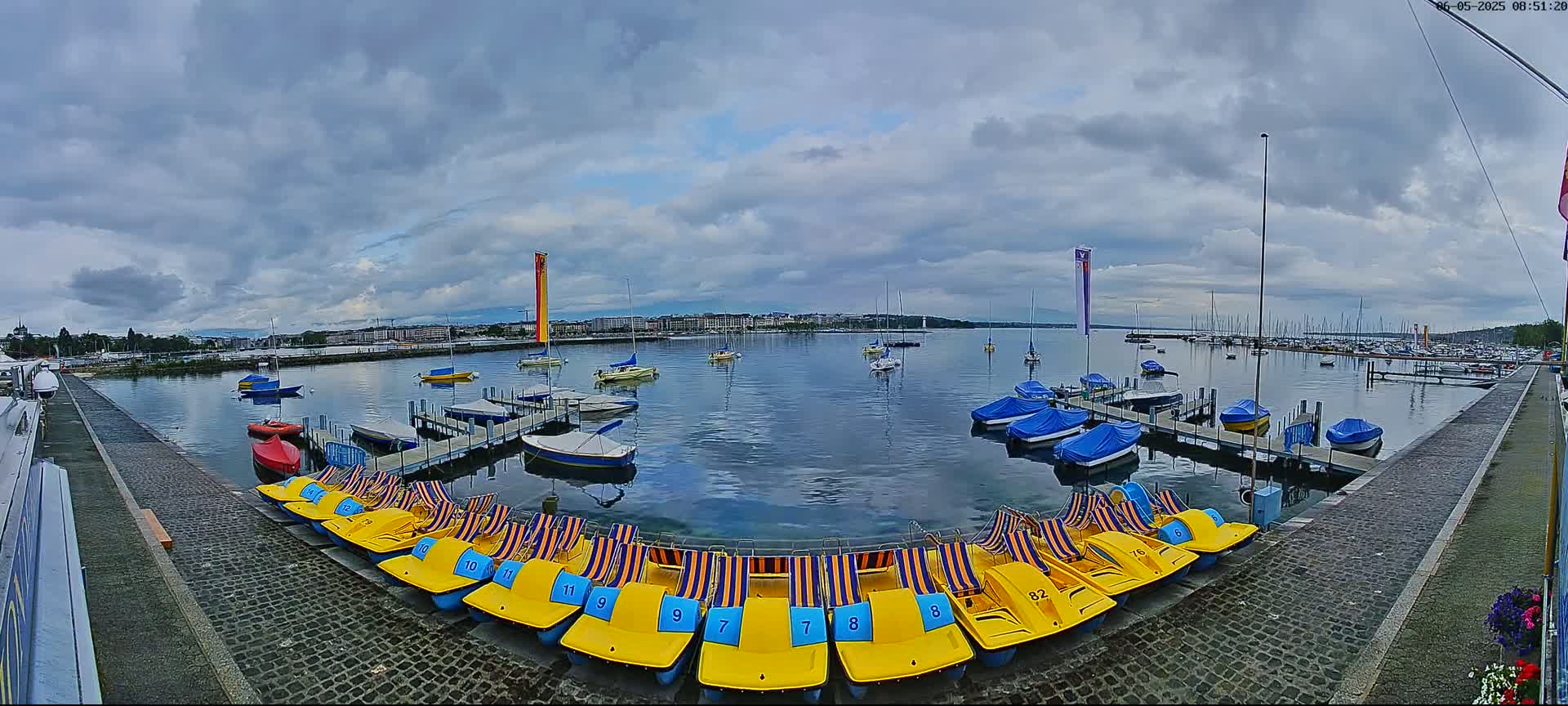 A calm lake with numerous boats docked, many covered, under a cloudy sky, with yellow paddle boats lined up on a cobblestone path.