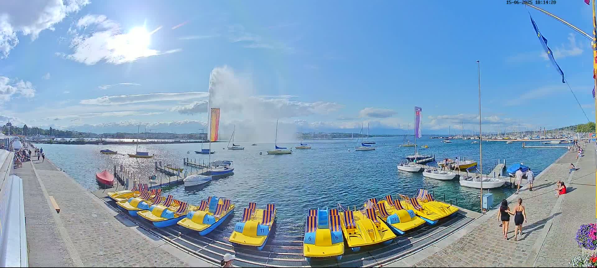 A sunny day on a lake shows a large water fountain, numerous boats docked and afloat, and people walking along a paved walkway.