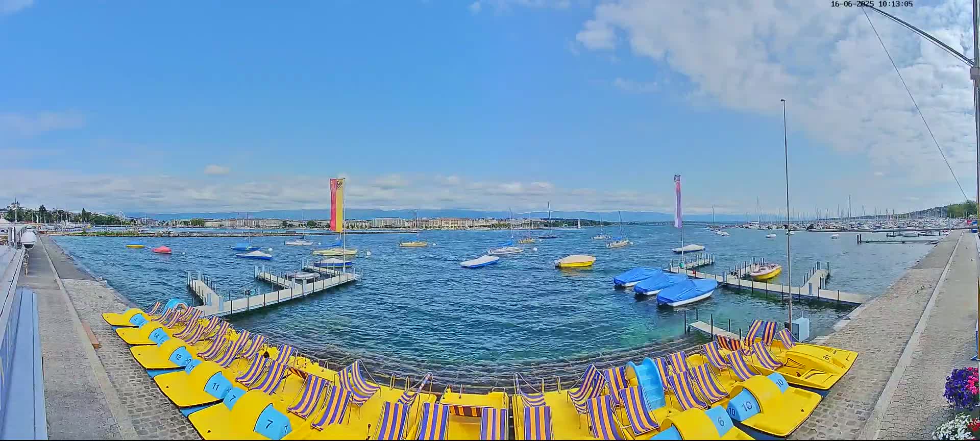A sunny day shows a calm body of water with numerous small boats and paddle boats docked at a shore lined with a paved walkway.