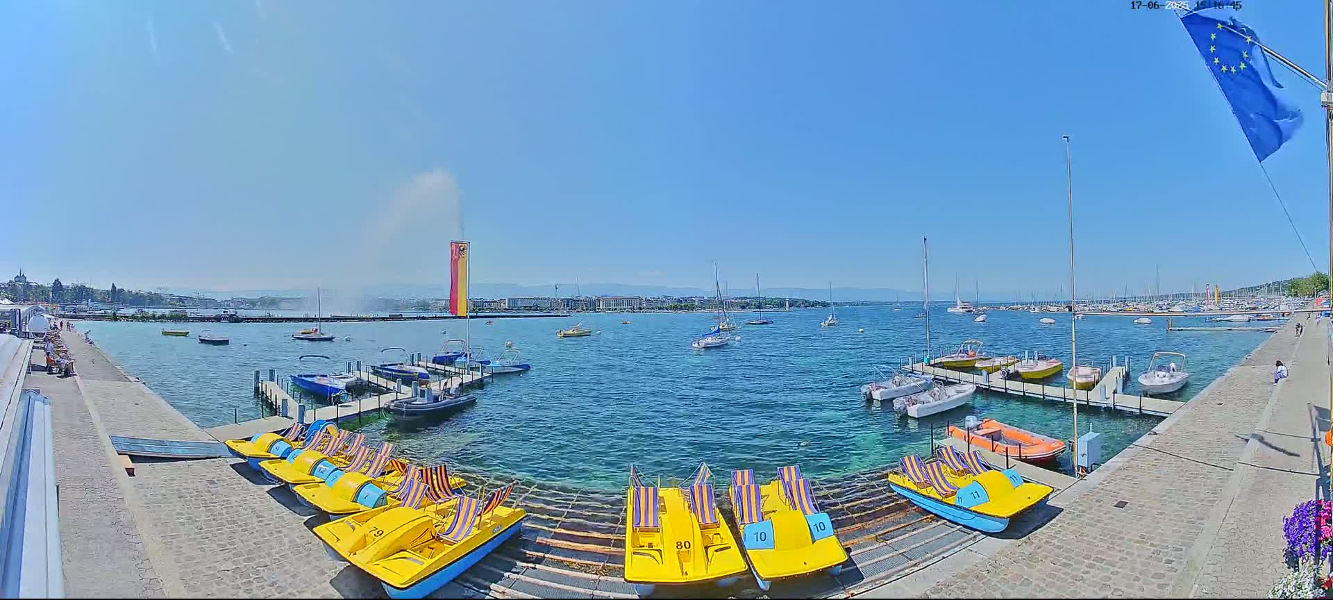 A sunny day shows a harbor filled with various boats and pedal boats, with a large water fountain spraying in the distance.