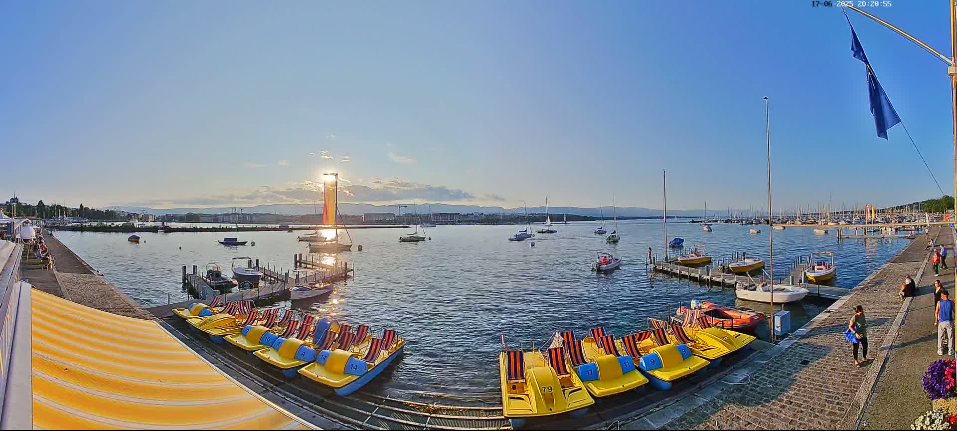 Under a clear, sunny sky, a calm lake is filled with numerous boats and paddle boats docked near a paved walkway with people strolling by.