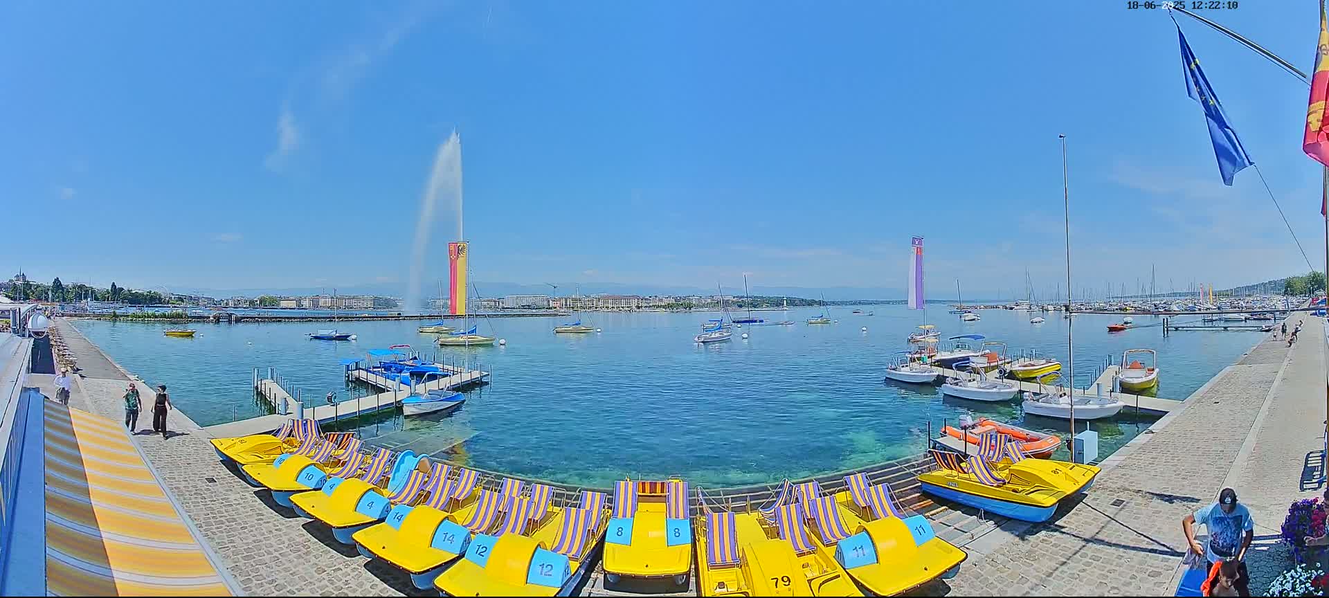 A waterfront scene on a sunny day shows numerous yellow paddle boats docked along a stone walkway, with several other boats and two tall water fountains in the background across a calm body of water.