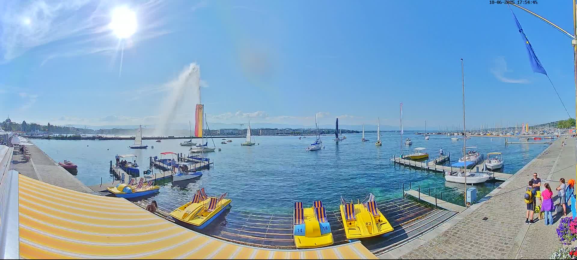 A sunny day shows a harbor filled with boats of various sizes, including pedal boats, near a tall water fountain spraying water high into the air.