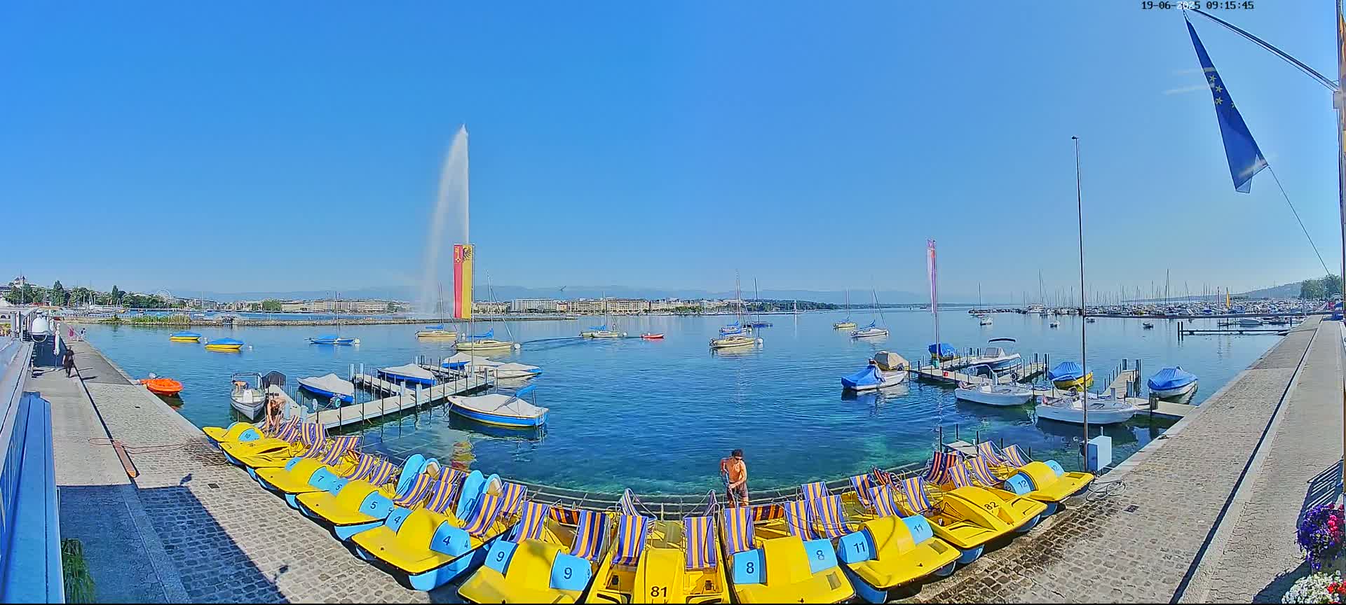 Under a clear blue sky, a large water fountain sprays high into the air over a calm lake, where numerous boats are docked near a row of paddle boats.