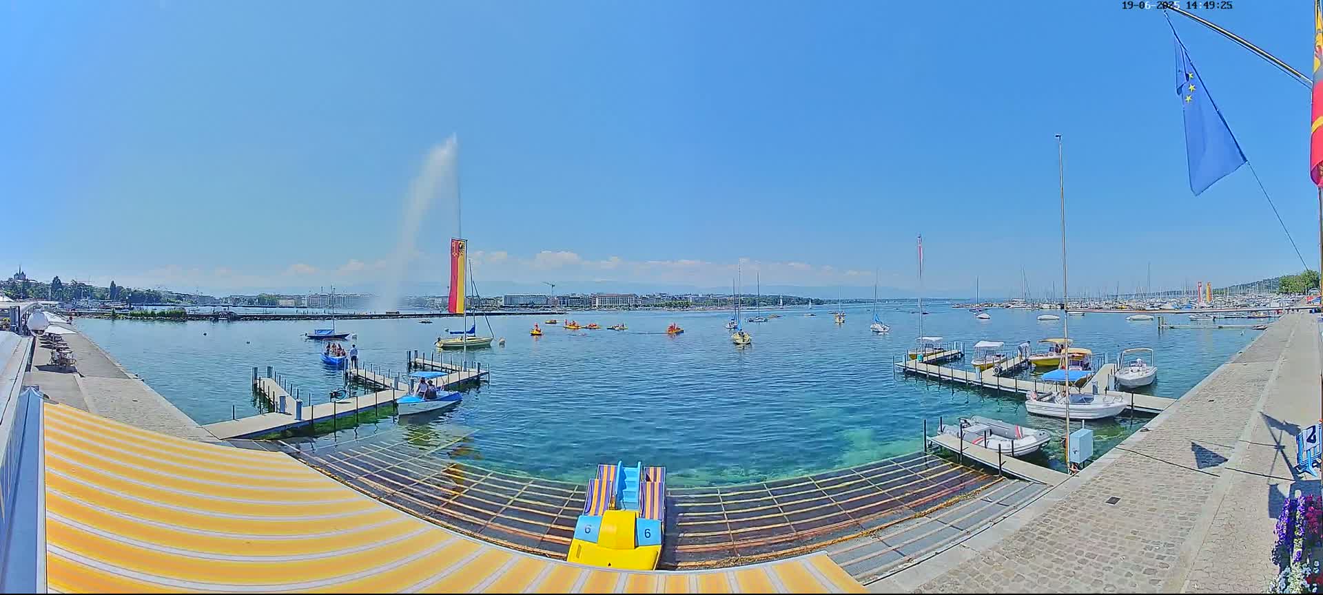 A clear, sunny day at a harbor shows calm, teal water with numerous boats docked and some in the water, a tall water fountain in the background, and a striped awning in the foreground.