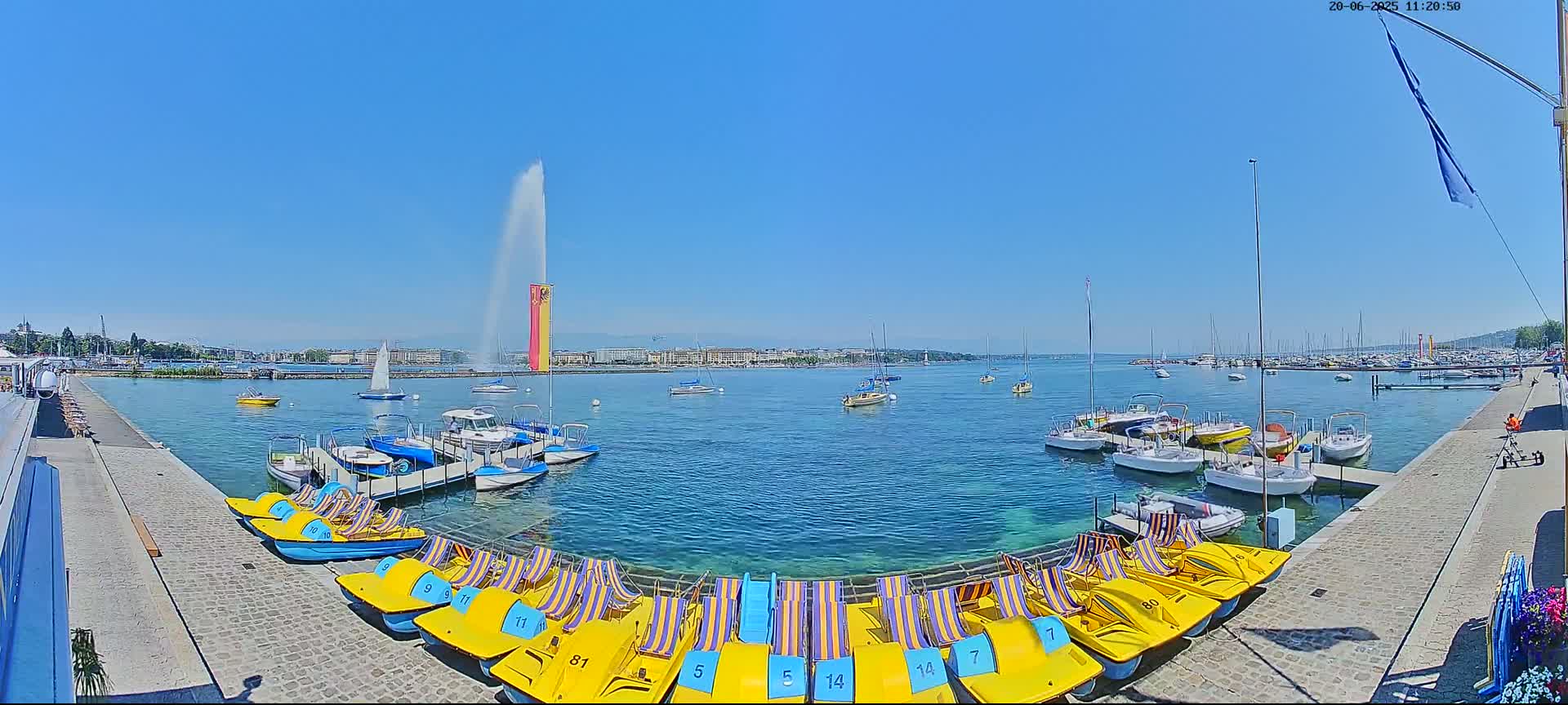 A sunny day on a lake with a large fountain in the background, numerous boats docked, and several paddle boats lined up on a stone embankment.