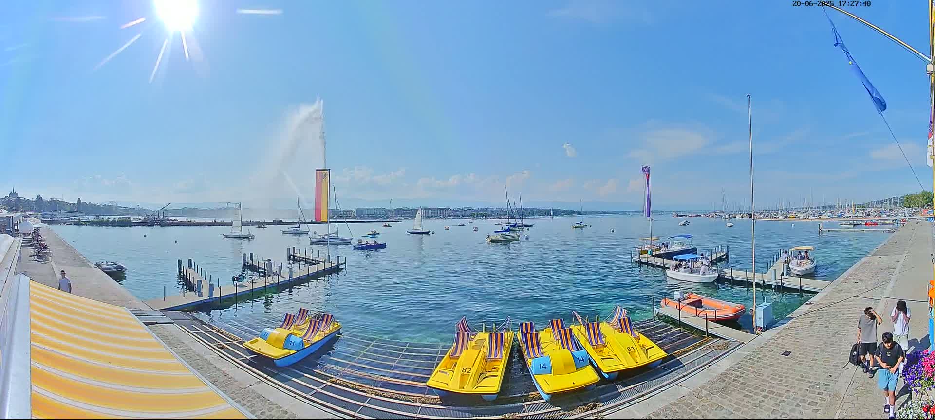 A sunny day on a lake shows numerous boats and paddle boats docked near a shoreline, with a large water fountain spraying water in the background.