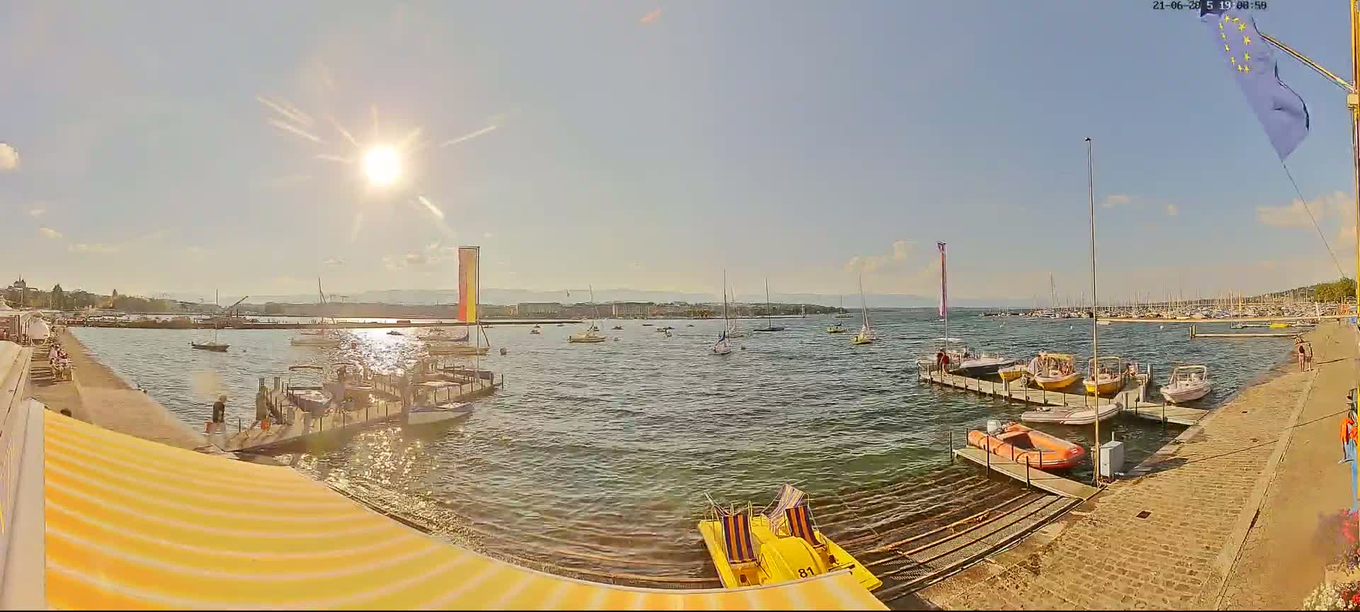 A sunny day at a harbor shows numerous boats docked and on the water, with a distant shoreline visible under a mostly clear sky.