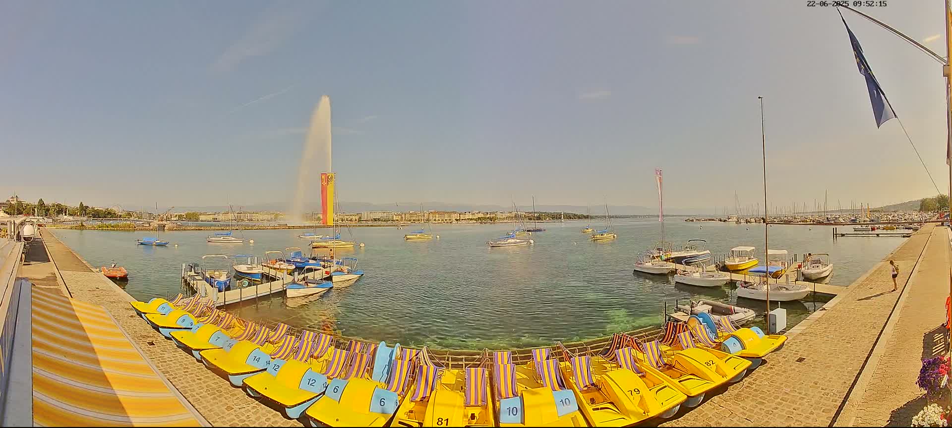 A large fountain sprays water high into a sunny sky over a calm lake with numerous boats docked along a paved waterfront.