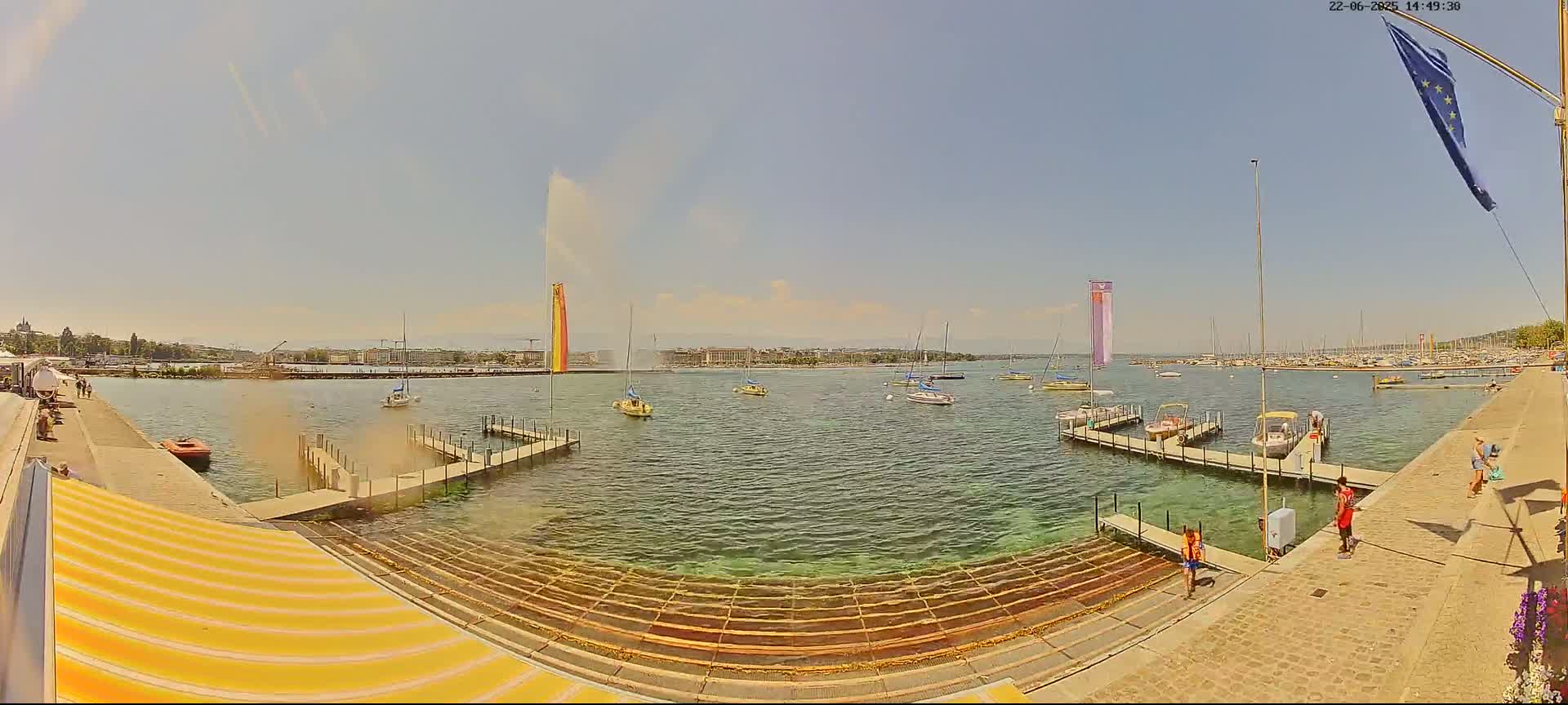 A large water fountain sprays water high into the air over a calm lake with several boats and docks on a sunny day.