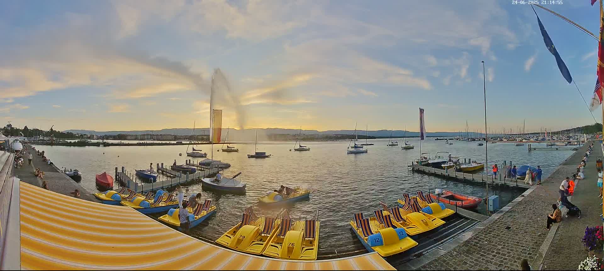 A large water fountain sprays water high into the air over a calm lake at sunset, with numerous small boats and pedal boats docked nearby, and people walking along a stone path.