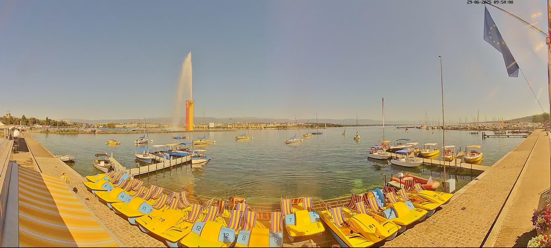 A large fountain sprays water into a sunny sky over a calm lake with numerous boats and paddle boats docked at a stone quay.