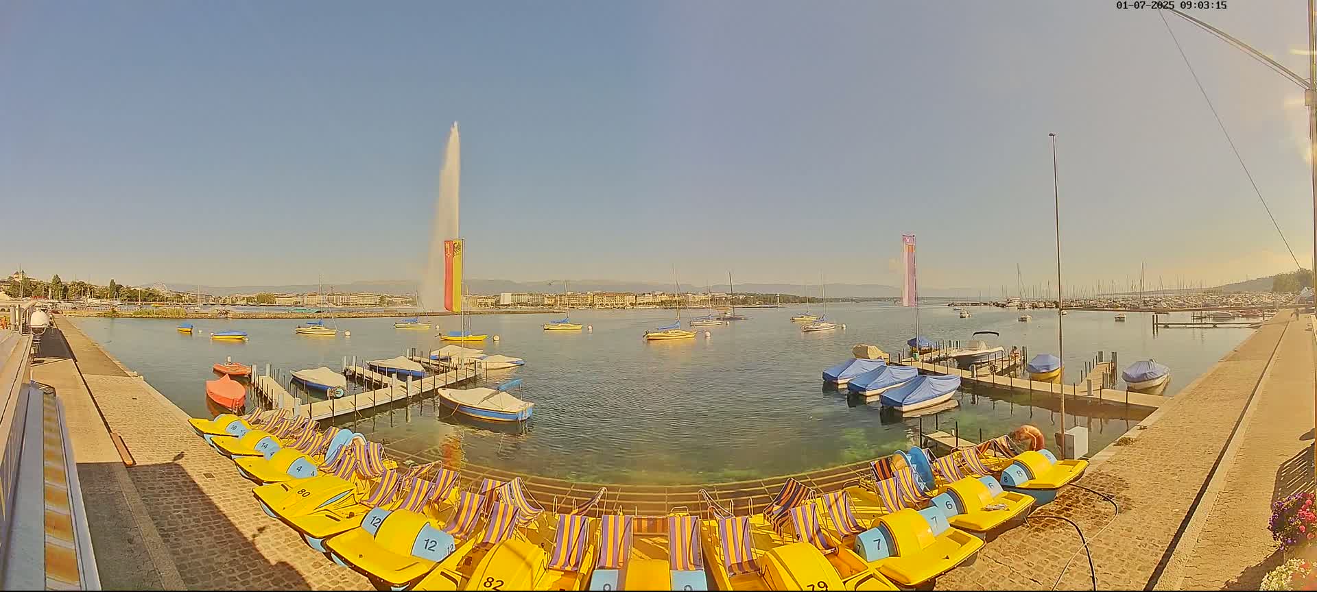 Under a clear blue sky, a lakefront scene shows numerous small boats moored at docks, pedal boats lined up on a paved walkway, and a large fountain spouting water in the distance.