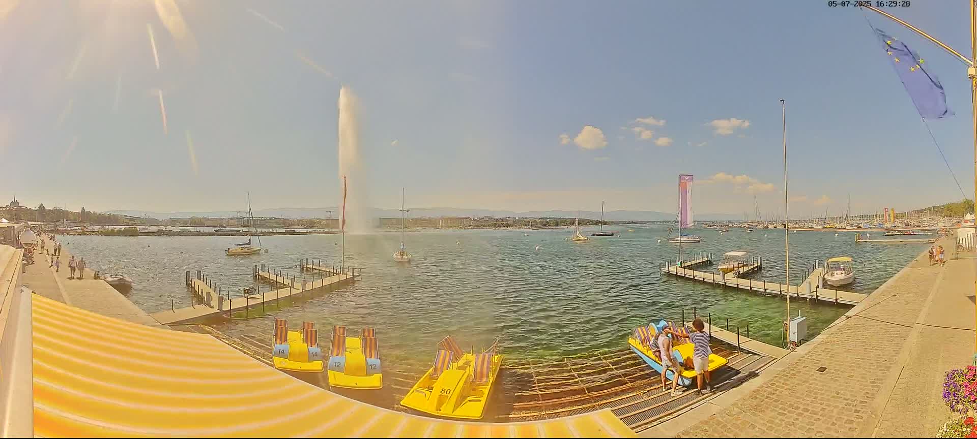 A sunny day shows a lake with a tall fountain spraying water, several boats, and people on a dock with paddle boats.