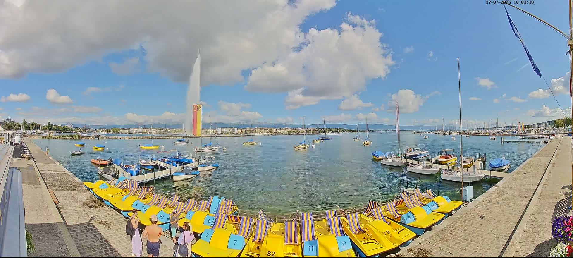 A large water fountain sprays water high into a partly cloudy sky over a calm lake with numerous docked boats and paddle boats, and a few people are standing near the paddle boats on a stone walkway.