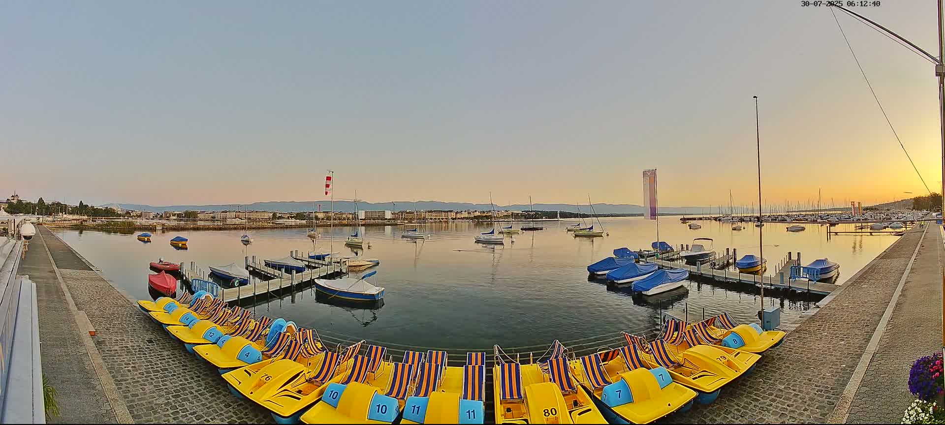 A large water fountain sprays water high into a partly cloudy sky over a calm lake with numerous docked boats and paddle boats, and a few people are standing near the paddle boats on a stone walkway.