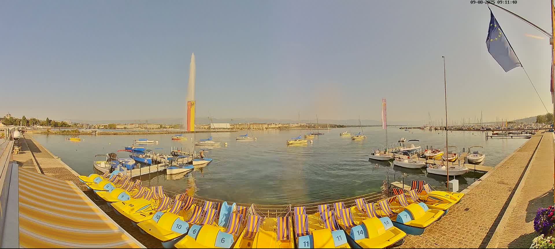 Under a clear blue sky, numerous boats are moored at a harbor next to a row of yellow paddle boats, with a tall fountain featuring a vertical banner in the background.