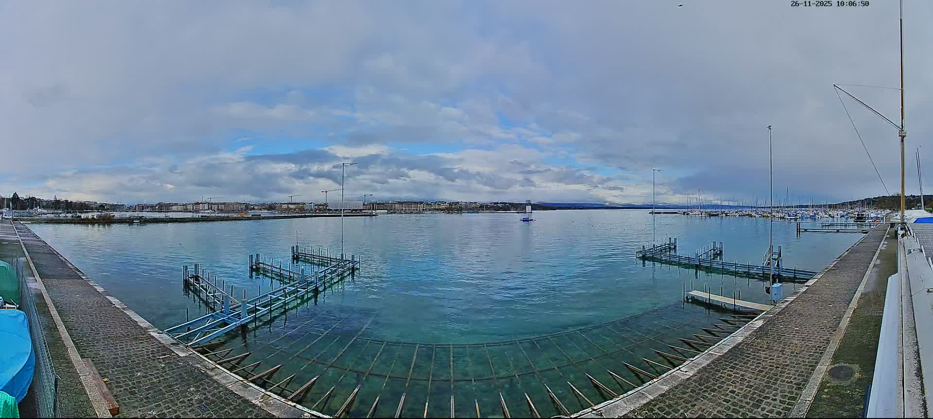 A panoramic view shows a marina with docks and boats on a clear lake, bordered by a distant cityscape and snow-capped mountains under a partly cloudy sky.