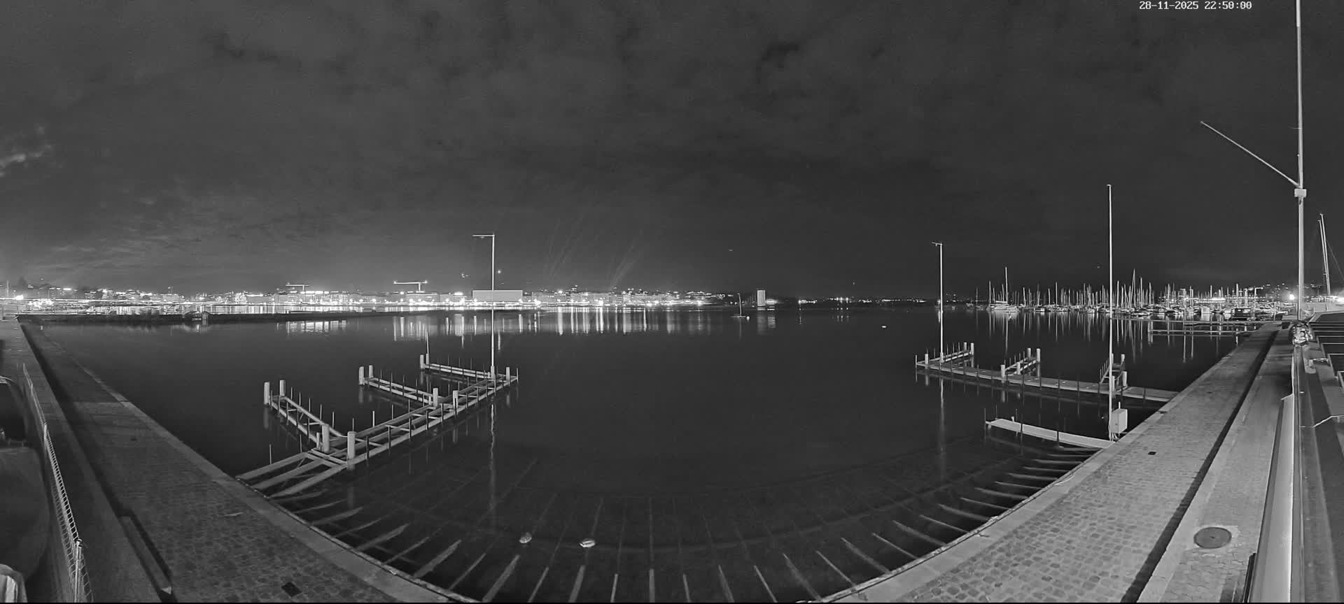 Under a cloudy night sky, a grayscale panoramic view captures a calm harbor with city lights reflecting on the distant water, numerous boats moored on the right, and empty piers in the foreground.