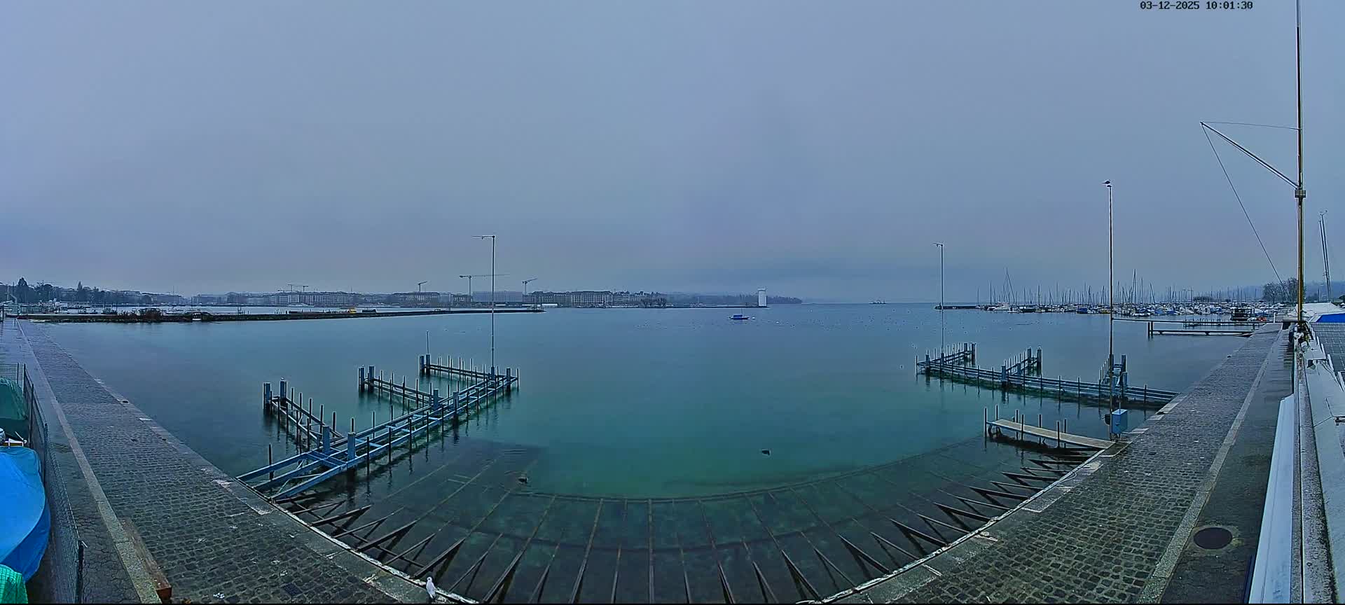 On an overcast day, a wide-angle view captures a marina with foreground docks, numerous sailboats, and a distant city skyline with a prominent white structure across the expansive, calm water.