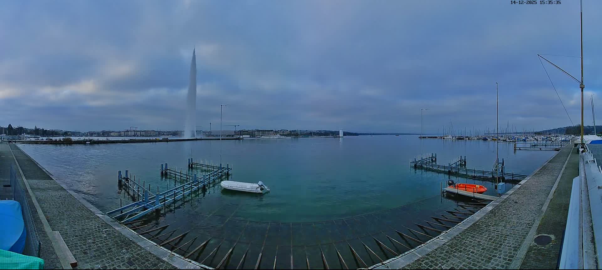 On an overcast day, a wide-angle view captures a marina with foreground docks, numerous sailboats, and a distant city skyline with a prominent white structure across the expansive, calm water.