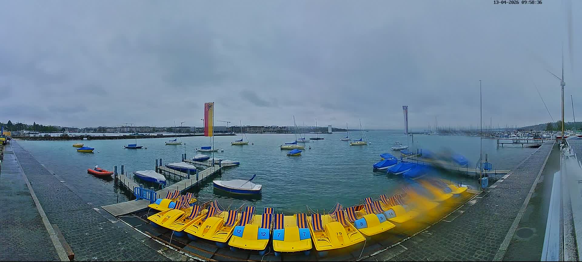 On an overcast day, a wide-angle view captures a marina with foreground docks, numerous sailboats, and a distant city skyline with a prominent white structure across the expansive, calm water.