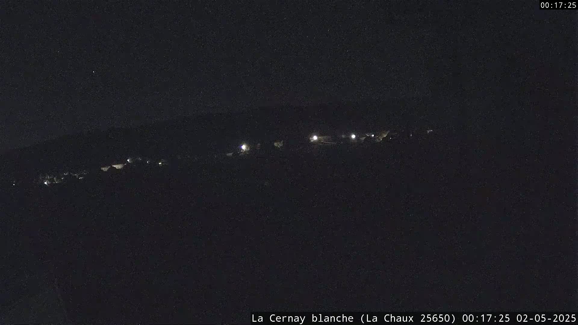 A nighttime long shot shows a line of faintly lit buildings on a dark hillside under a clear, starless sky.