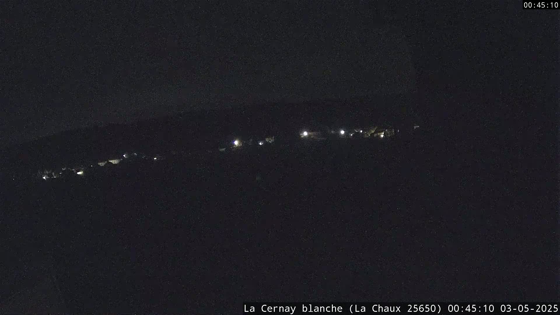 A nighttime, long shot shows a dark landscape with a few faint lights visible in the distance.