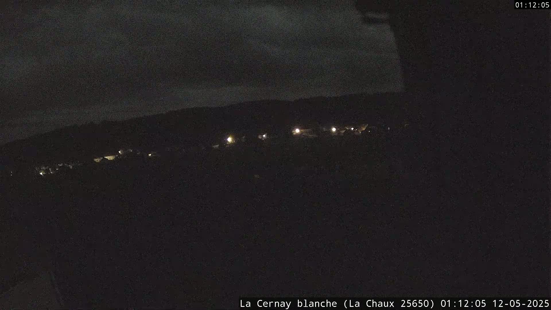 A nighttime, long-range view shows a dark landscape with a few faint lights in the distance under a cloudy sky.