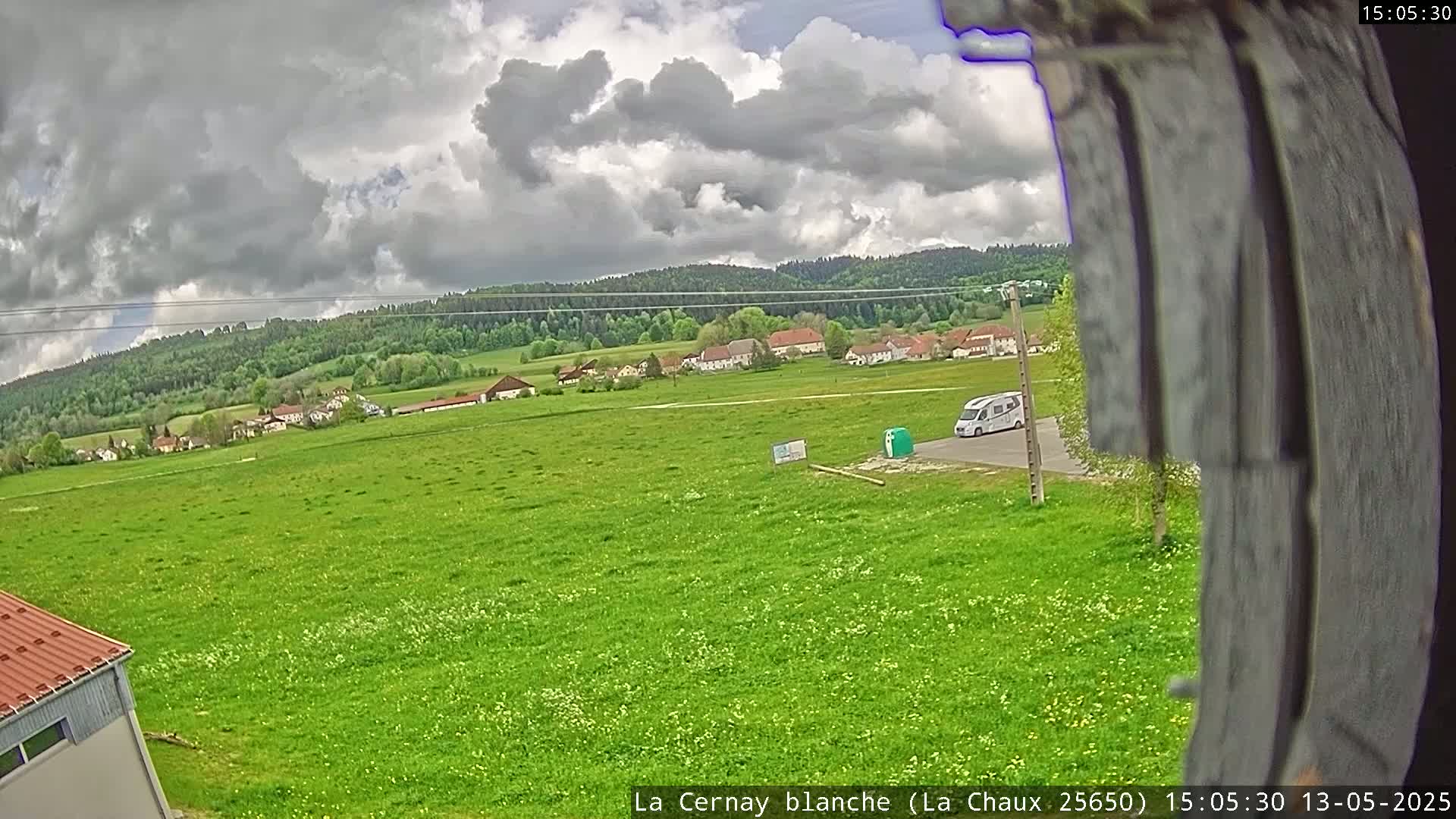 A large green field stretches to a small village nestled amongst green hills under a cloudy sky.
