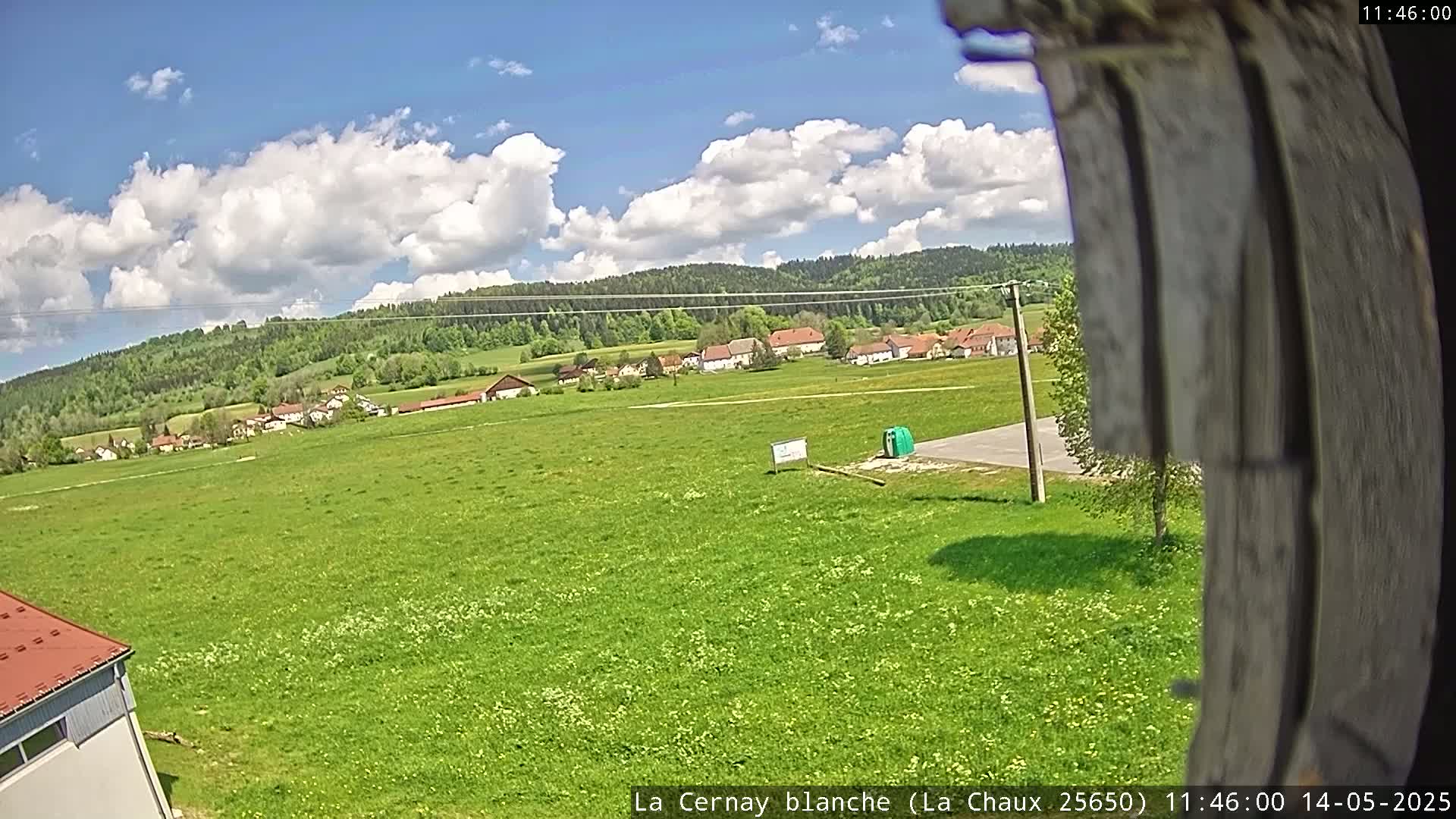 A partly cloudy, sunny day reveals a green field with a small village and forest in the background.