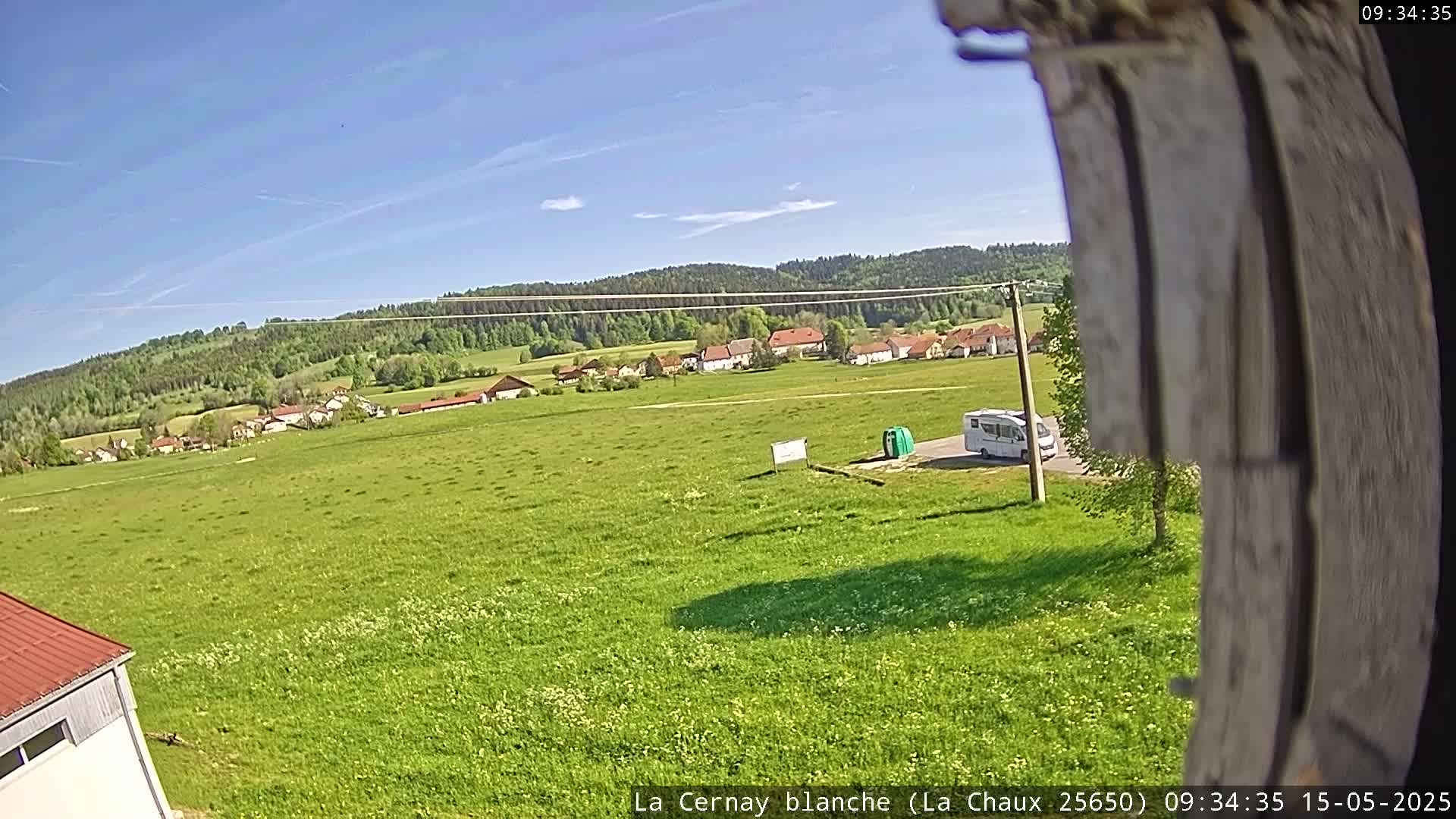 A camper van is parked in a grassy field, overlooking a village nestled in a valley surrounded by a forest under a clear blue sky.