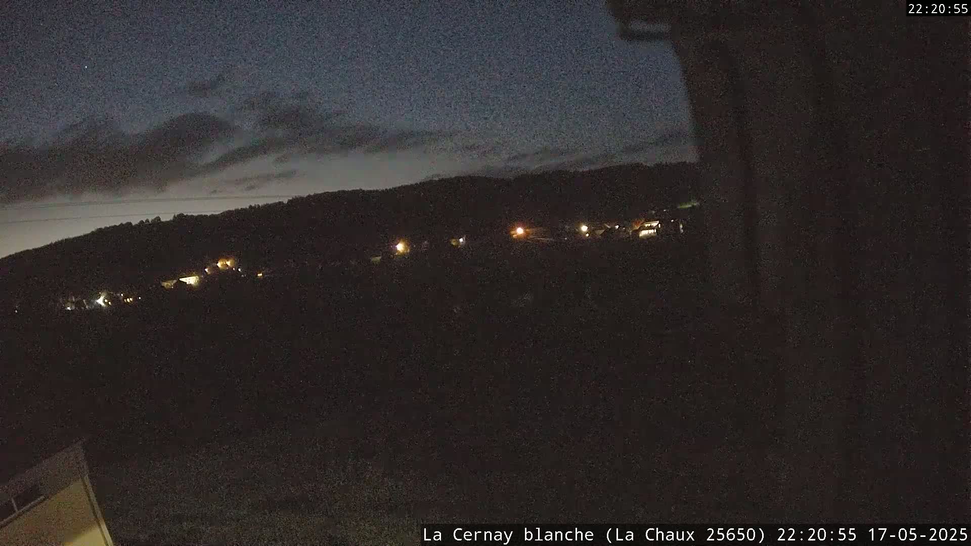 A nighttime view of a dark hillside with a few scattered lights visible under a mostly cloudy sky.