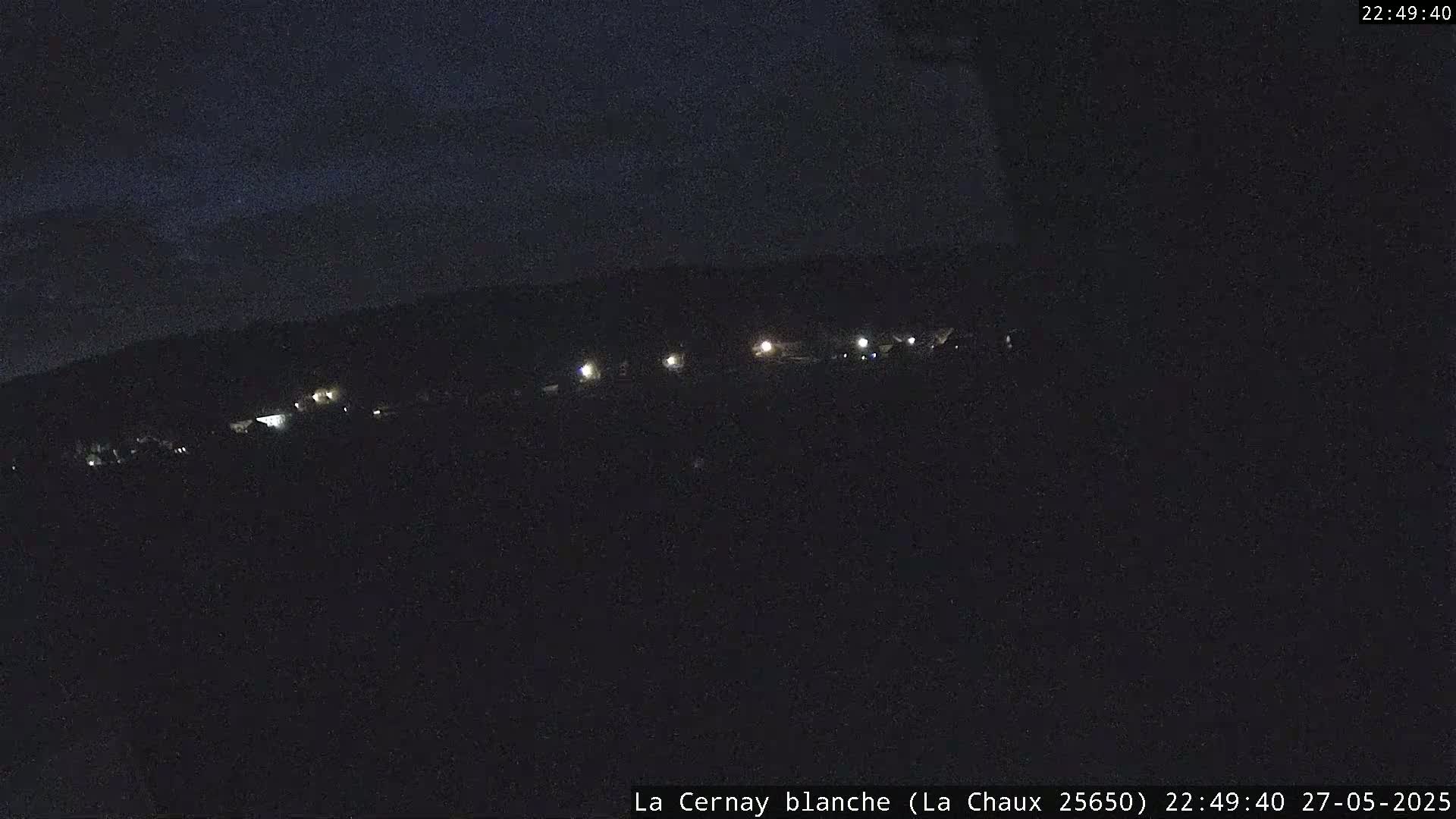 A nighttime, low-light view shows a dark landscape with a line of faint lights in the distance under a dark sky.