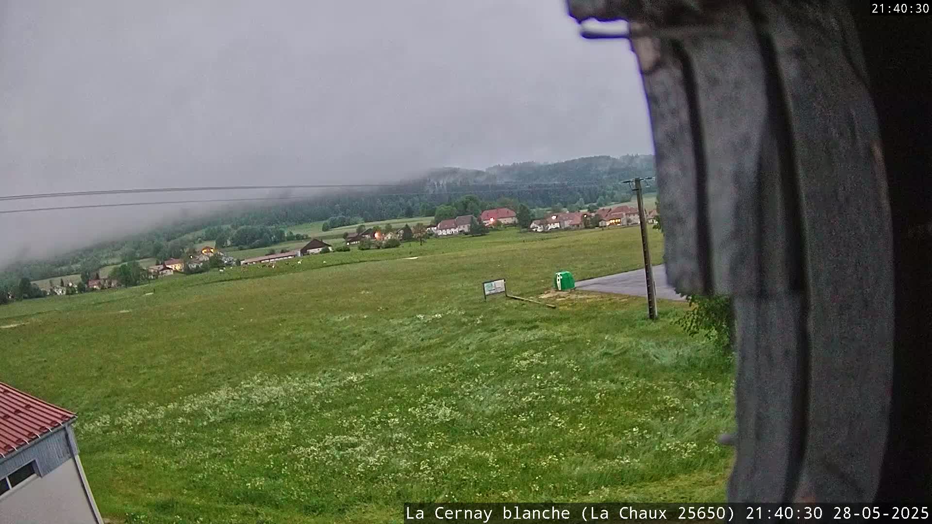A mostly overcast sky hangs over a green field, a small village, and a forest in the distance.