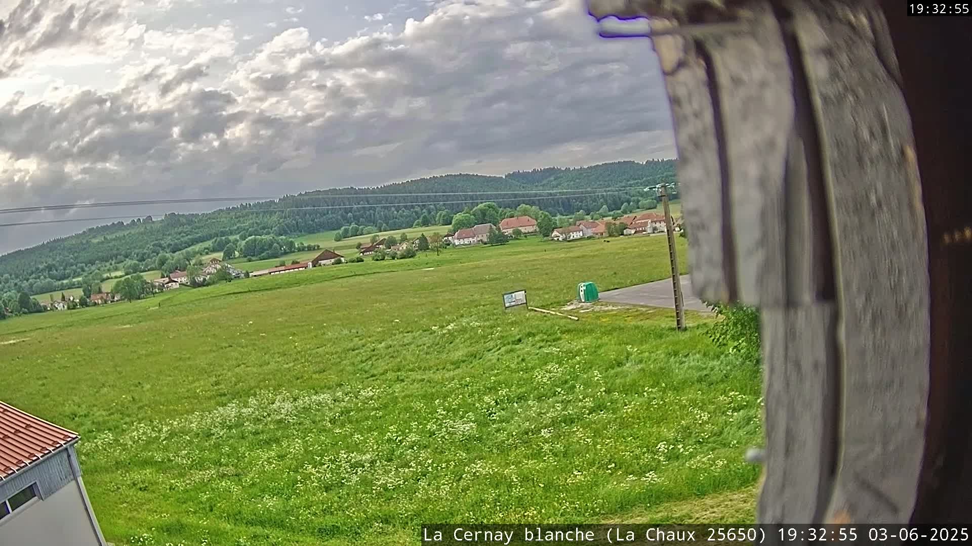 A mostly cloudy day reveals a wide expanse of green field with a small village and forest in the background.