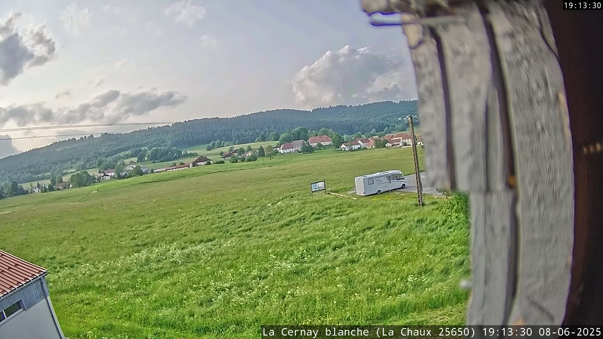 A white camper van is parked in a grassy field, with a small village and forested hills visible in the background under a partly cloudy sky.