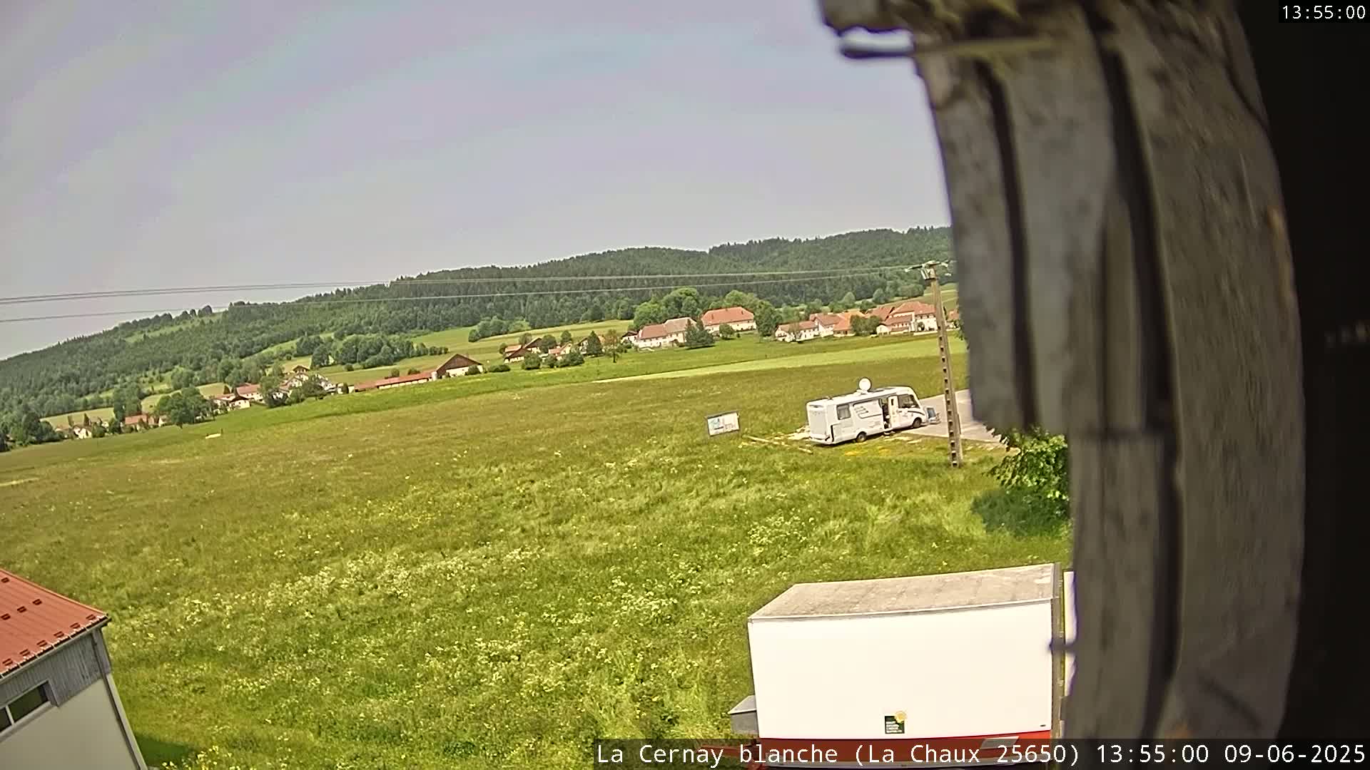 A large field with wildflowers, a small village in the distance, a camper van, and a white trailer are visible under a clear sky.